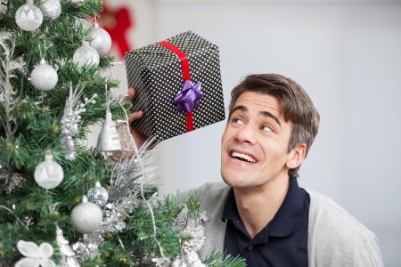 Curious mid adult man holding gift by Christmas tree at homeの写真素材