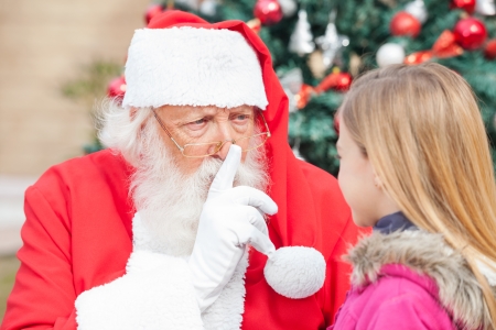 Santa Claus gesturing finger on lips while looking at girl outdoorsの写真素材