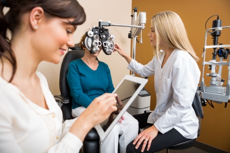 Female optometrist examining senior woman's eyes while colleague using digital tablet in store