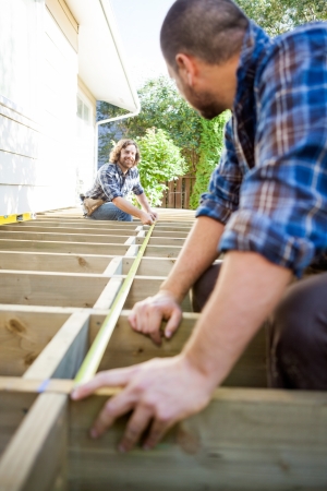 Mid adult carpenter measuring wood with tape while looking at coworker assisting himの写真素材