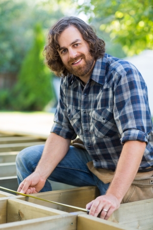 Portrait of confident construction worker measuring wood with tape at construction siteの写真素材
