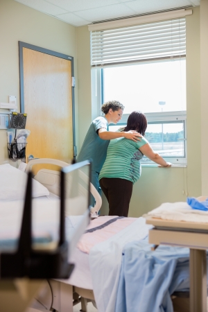 Female nurse consoling pregnant woman having a contraction and supporting herself by windowの写真素材