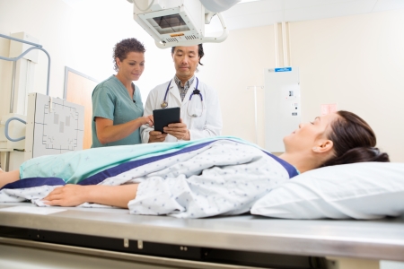 Mid adult nurse with doctor using digital tablet while patient lying on xray table in hospital roomの写真素材