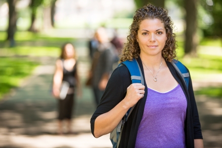 Portrait of confident female grad student with backpack standing at campus with friends in backgroundの写真素材