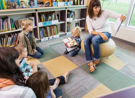 Young teacher reading book to children in library of elementary school
