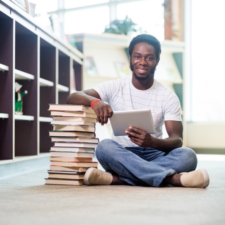 Full length portrait of young male student with stacked books and digital tablet sitting on floor at libraryの写真素材