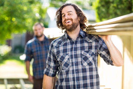 Mid adult carpenter with coworker carrying wooden planks while laughing outdoorsの写真素材