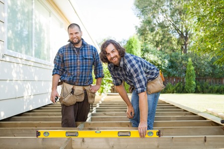 Portrait of happy construction worker checking level of wood with spirit level while standing with...