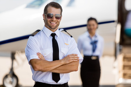 Portrait of confident pilot with arms crossed against stewardess and private jet at terminal