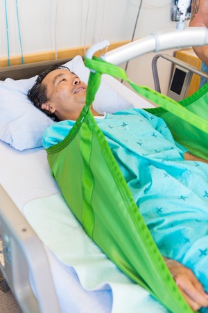 Mature male patient lying on sling of hydraulic lift on bed at hospitalの写真素材