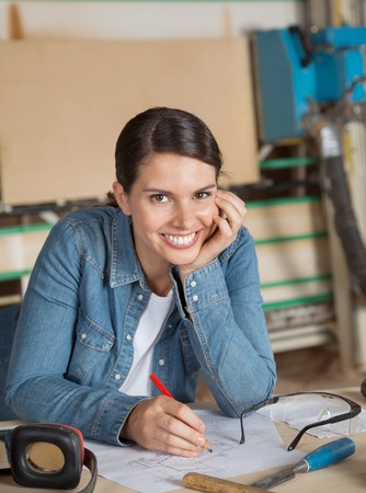 Portrait of happy female carpenter with hand on chin working on blueprint table in workshopの写真素材
