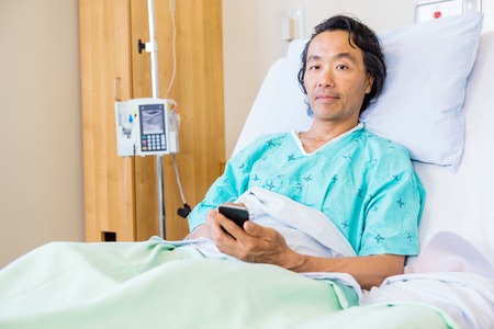 Patient Holding Mobile Phone While Resting On Hospital Bedの写真素材
