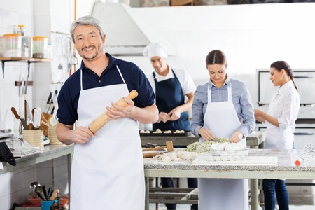 Confident Male Chef Holding Rolling Pin While Colleagues Prepariの写真素材