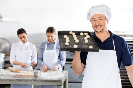 Smiling Chef Carrying Baking Sheet With Dough Balls At Kitchen