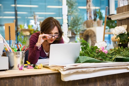Shocked mid adult female florist holding glasses while using laptop at counter in flower shopの写真素材