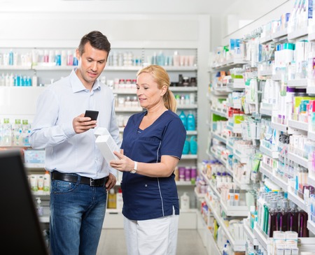 Male customer using mobile phone while chemist holding products in pharmacyの写真素材
