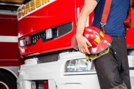 Midsection of young fireman holding red helmet while leaning on firetruck at stationの写真素材