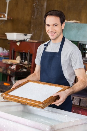 Portrait of confident mid adult worker dipping mold in pulp and water at paper factoryの写真素材