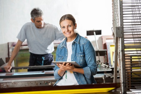 Portrait of smiling female worker holding digital tablet while colleague working in background at...