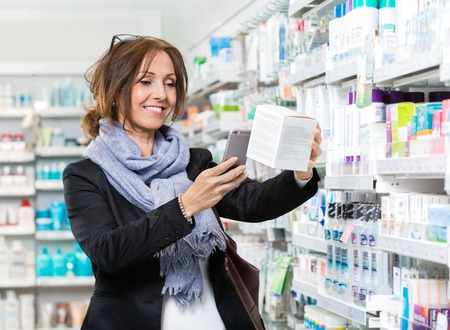Smiling female customer scanning product through mobile phone in pharmacyの写真素材