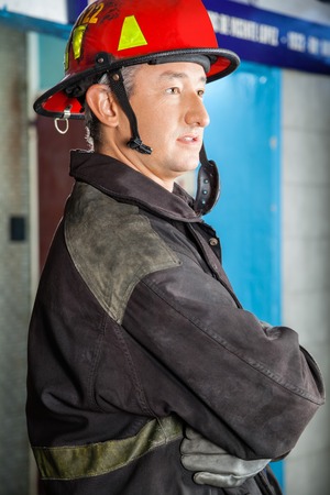 Side view of confident firefighter in uniform standing arms crossed at fire stationの写真素材