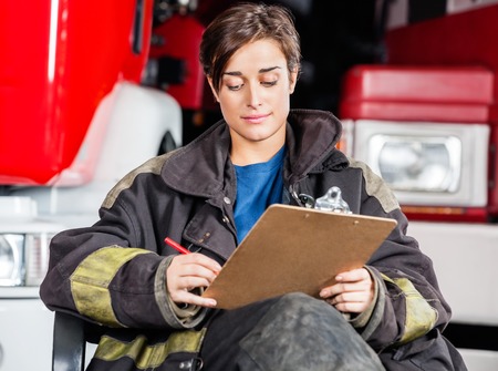 Young firewoman writing on clipboard while sitting against trucks at fire station