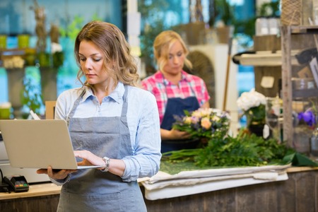 Female florist using laptop with colleague in background at flower shopの写真素材