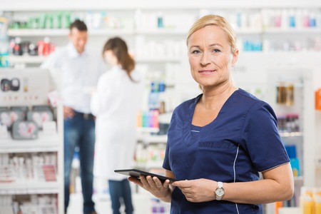 Portrait of confident assistant holding digital tablet while pharmacist and customer standing in background at pharmacyの写真素材