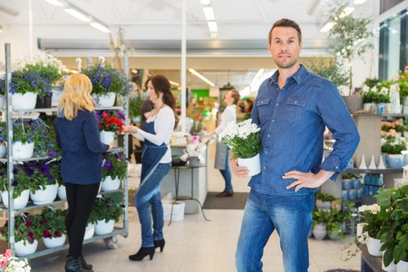 Portrait of confident man holding pot plant with florist assisting customer in background at shopの写真素材