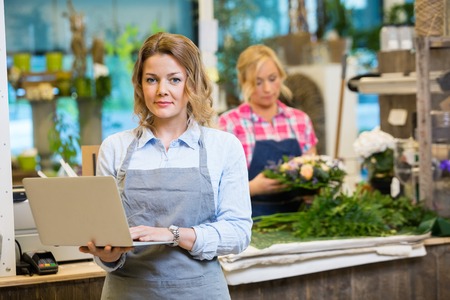 Portrait of female florist using laptop with colleague working in background at flower shopの写真素材