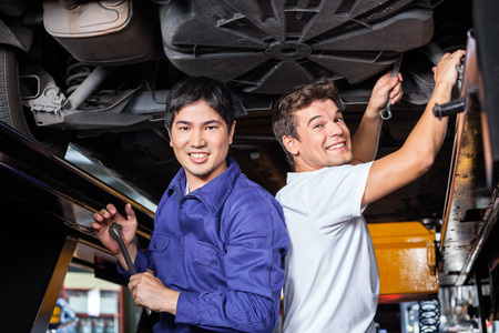 Portrait of happy male mechanics working under lifted car at auto repair shopの写真素材
