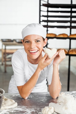 Portrait of beautiful baker holding heart shape dough while leaning on table in bakeryの写真素材
