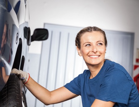 Happy female mechanic looking away while fixing car tire at auto repair shopの写真素材