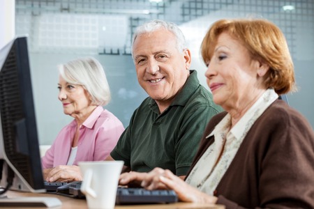 Portrait of confident senior man sitting in computer class with female friends at deskの写真素材