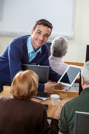 Portrait of confident teacher showing digital tablet to senior students at desk in computer class