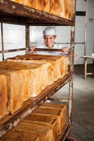 Smiling female baker pushing rack full of freshly baked breads in bakeryの写真素材