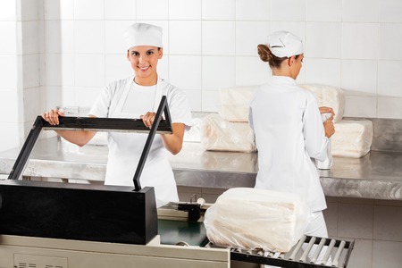 Portrait of confident female baker using vacuum seal machine while coworker working in bakery