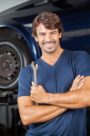 Portrait of smiling mechanic holding wrench while standing arms crossed at repair shopの写真素材