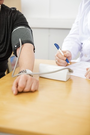 Cropped image of doctor holding pen while examining female patient's blood pressure at table in...