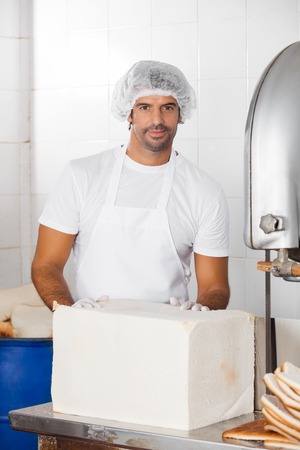 Portrait of confident male baker with bread loaf at cutting machine in bakery