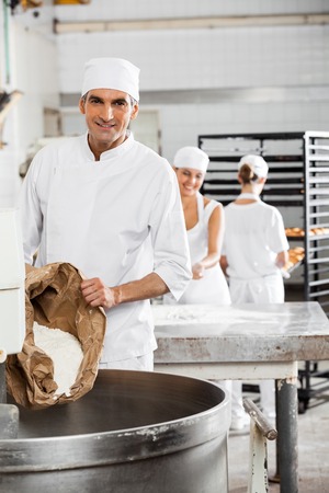 Portrait of smiling mature baker pouring flour in kneading machine at bakeryの写真素材