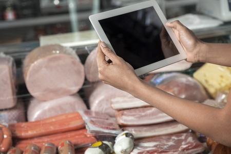 Cropped image of young woman photographing cheese and sausages through tablet computer in grocery storeの写真素材