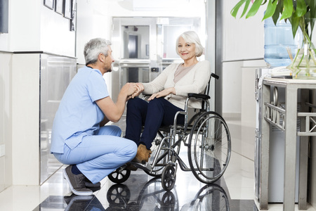 Mature male physiotherapist holding smiling senior woman's hand on wheelchair in rehab center