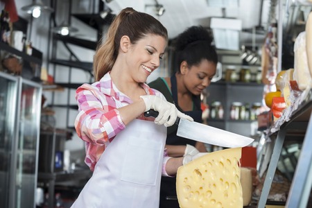 Happy young woman slicing cheese at counter with colleague working in backgroundの写真素材