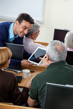 Senior male student asking question to teacher on digital tablet at desk in computer class