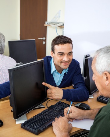 Happy male teacher showing something to senior student on computer at desk in classroom