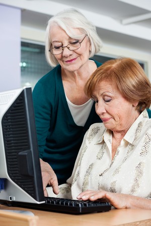 Happy senior woman assisting female friend in using computer at classroomの写真素材