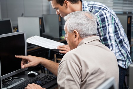 Male teacher holding clipboard while assisting senior man in using computer at classroomの写真素材