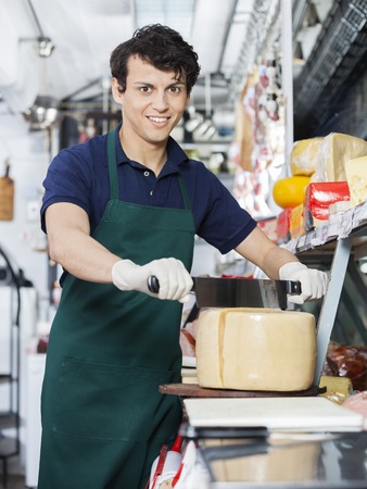 Portrait of happy young salesman slicing whole cheese with double handled knife at counter in shopの写真素材