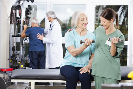 Smiling female nurse guiding senior woman exercising with dumbbell at rehab fitness center